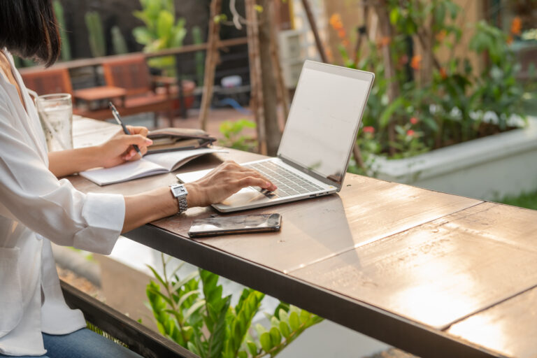 Femme travaillant sur ordinateur portable avec carnet et stylo en terrasse, évoquant la rédaction web, la correction et la relecture à distance.
